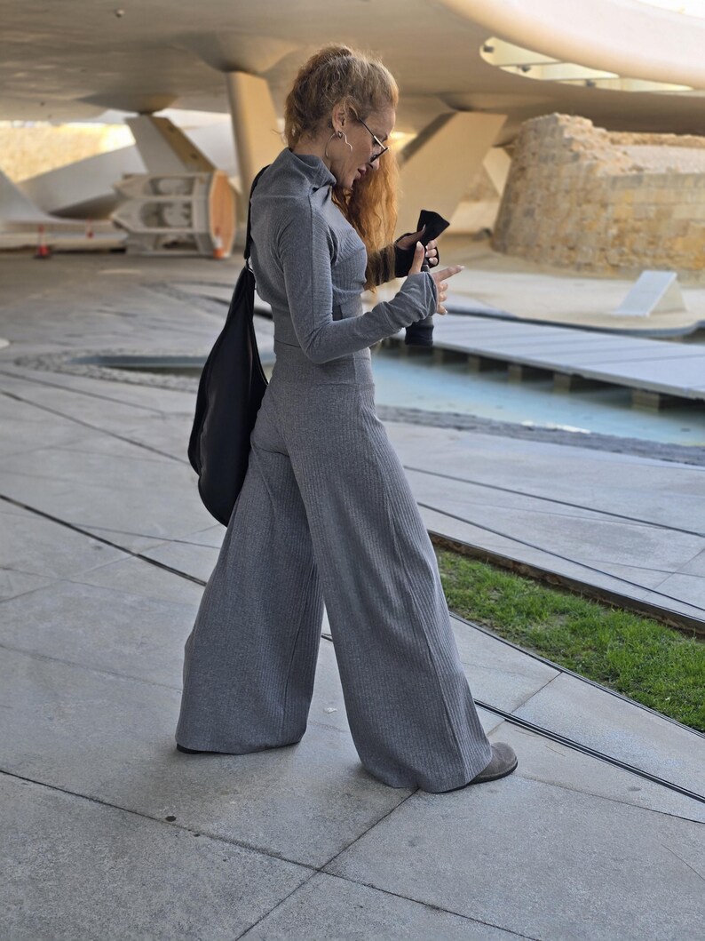 May include: A woman wearing a gray ribbed turtleneck top and matching wide-leg pants. She is standing on a concrete surface with a black bag slung over her shoulder.