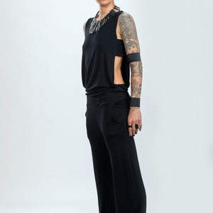 Könnte beinhalten: Eine Frau in einem schwarzen, ärmellosen Jumpsuit mit seitlichem Schlitz und einer schwarzen Halskette. Sie hat Tätowierungen auf ihren Armen und trägt schwarze Armbänder. Sie steht vor einem weißen Hintergrund.