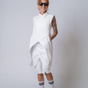 Könnte beinhalten: Eine ärmellose weiße Tunika mit Wickeloptik und überlappenden Stoffbahnen, kombiniert mit weißen knielangen Shorts. Das Outfit wird durch weiße Socken mit drei schwarzen Streifen und schwarze Sportschuhe mit weißer Sohle ergänzt.