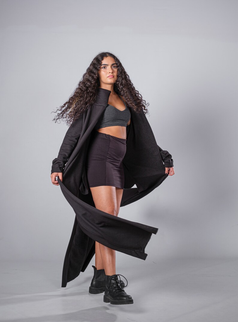 May include: A woman wearing a black long-sleeved cardigan with a flowing design, a black mini skirt, and black combat boots. Her hair is long and curly.