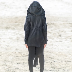 May include: A person wearing a black hooded sweatshirt with a long, flowing back, black leggings, and gray sneakers. The person is standing on a sandy surface.