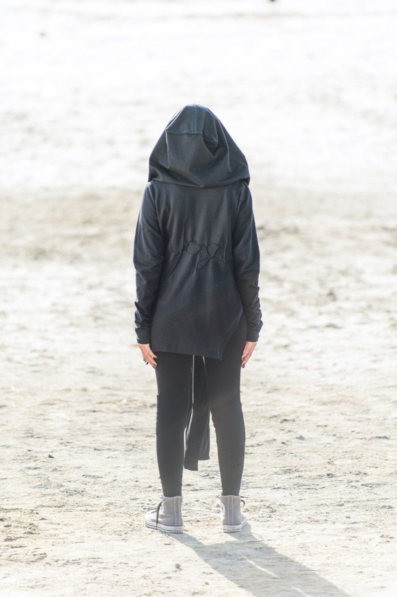 May include: A person wearing a black hooded sweatshirt with a long, flowing back, black leggings, and gray sneakers. The person is standing on a sandy surface.