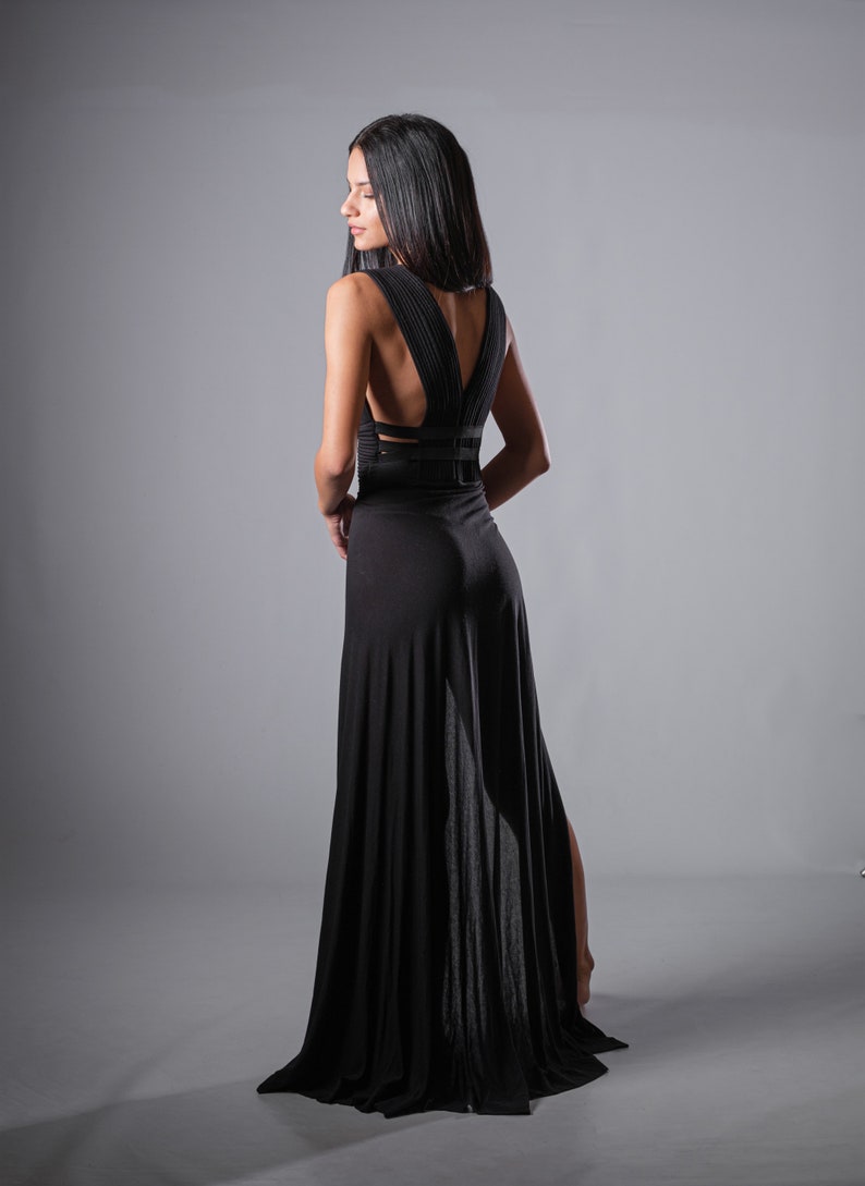 May include: A woman wearing a black maxi dress with a low back and a slit in the skirt. The dress has a criss-cross design on the back.