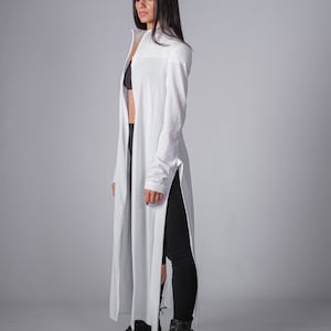 May include: A white long-sleeved shirt with a high neckline and side slits. The shirt is worn over a black bra and black leggings. The person is wearing black lace-up boots.