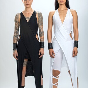 May include: Two people wearing long, flowing dresses. The person on the left is wearing a black dress with cutouts and has tattoos on their arms. The person on the right is wearing a white dress with a criss-cross design. Both people are wearing black wrist cuffs and black shoes.