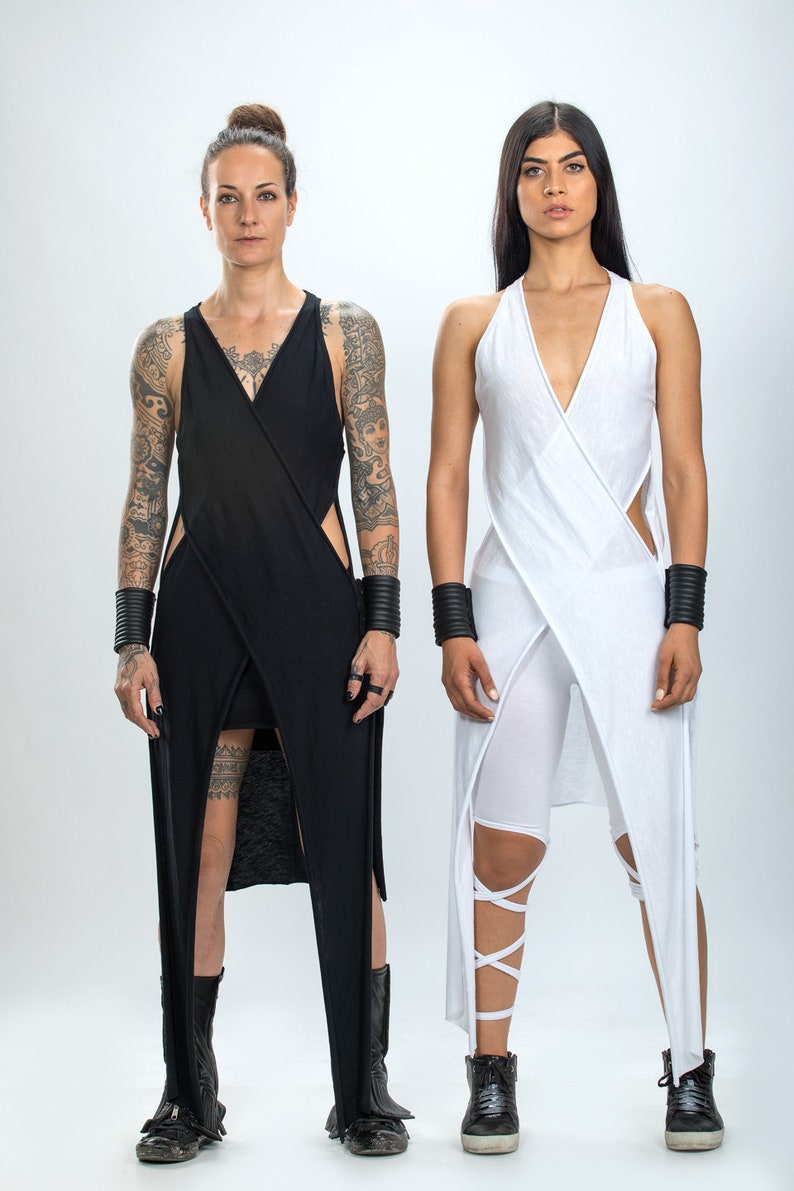 May include: Two people wearing long, flowing dresses. The person on the left is wearing a black dress with cutouts and has tattoos on their arms. The person on the right is wearing a white dress with a criss-cross design. Both people are wearing black wrist cuffs and black shoes.