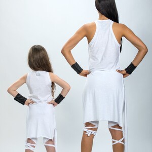 May include: Two people wearing white clothing with black wristbands. The person in the front is a child wearing a white tank top and white leggings with white straps. The person in the back is wearing a white tank top and white leggings with white straps. Both people are facing away from the camera.