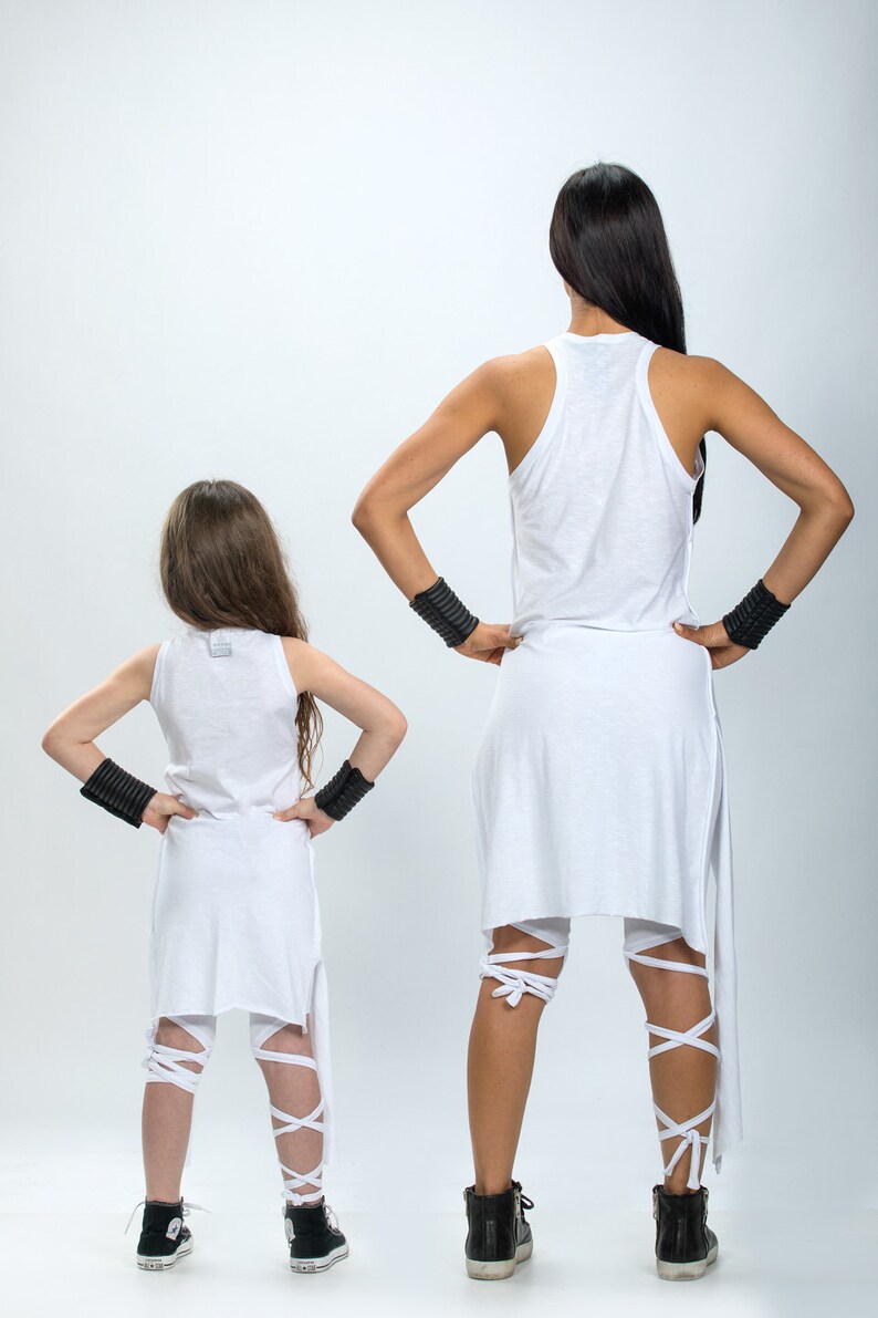 May include: Two people wearing white clothing with black wristbands. The person in the front is a child wearing a white tank top and white leggings with white straps. The person in the back is wearing a white tank top and white leggings with white straps. Both people are facing away from the camera.