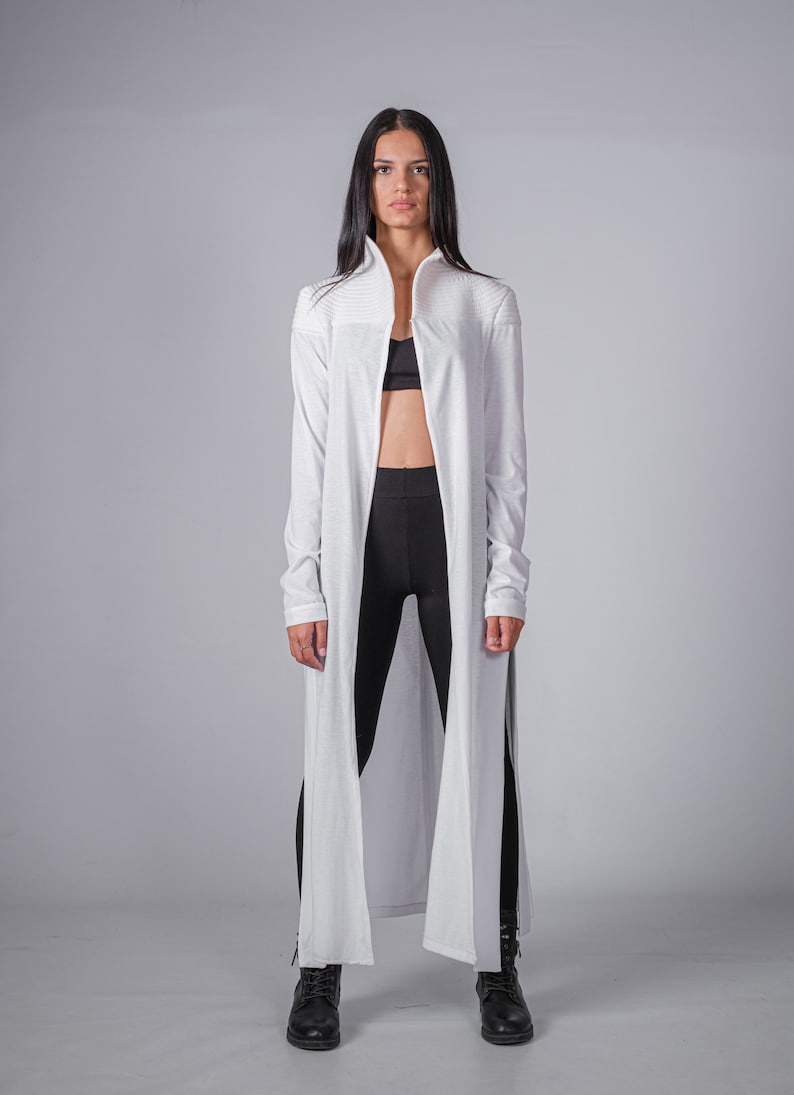 May include: A white long-sleeved cardigan with a high collar and a long, flowing design. The cardigan is open in the front, revealing a black sports bra and black leggings underneath.