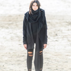 May include: A woman wearing a black hooded jacket with a long scarf, black leggings with cutouts, and black high-top sneakers. She is standing on a sandy surface.