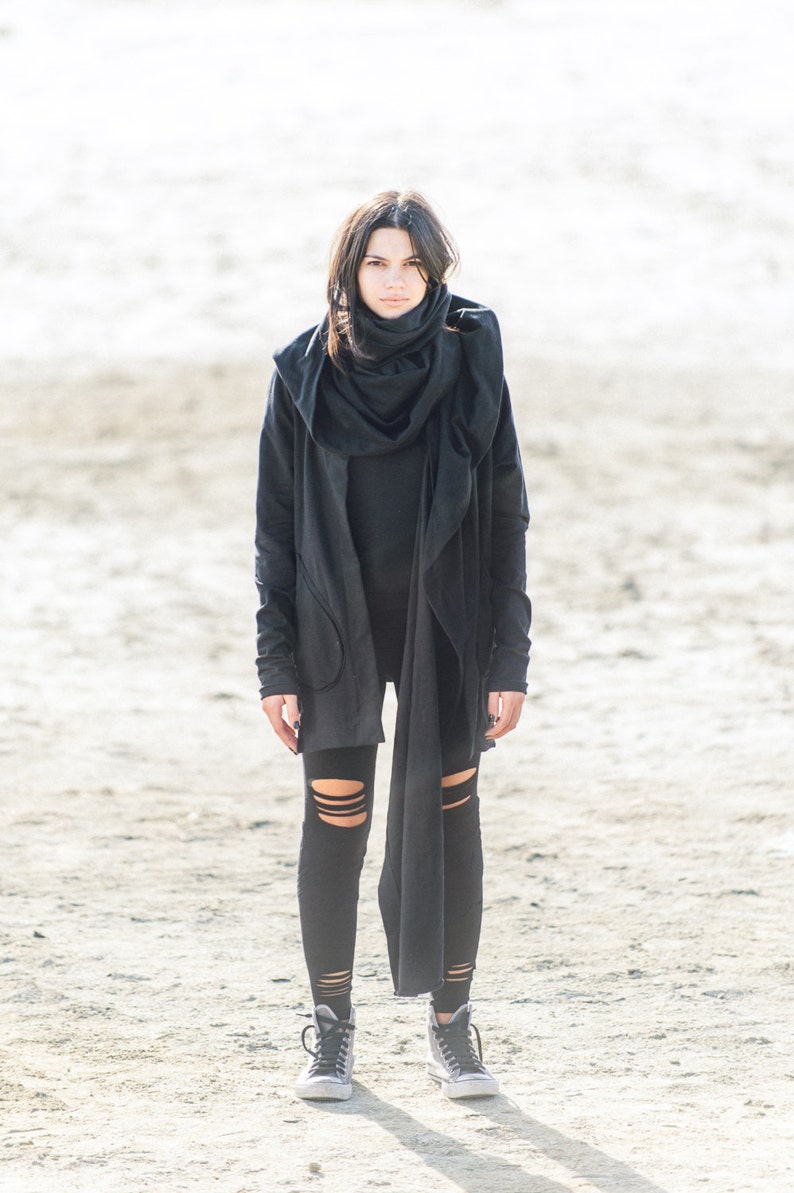 May include: A woman wearing a black hooded jacket with a long scarf, black leggings with cutouts, and black high-top sneakers. She is standing on a sandy surface.