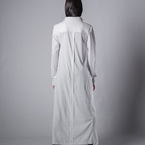 May include: A white long-sleeved maxi dress with a high neckline and a center seam down the back. The dress has a relaxed fit and a slightly flared skirt.
