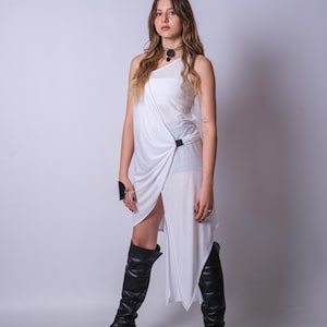 May include: A woman wearing a white one-shoulder dress with a black belt and black thigh-high boots. She is standing in front of a white background.