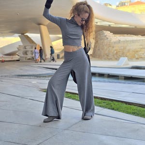 May include: A woman wearing a gray crop top and matching wide-leg pants. She is wearing black fingerless gloves and sunglasses. She is standing in front of a modern building with a curved roof.