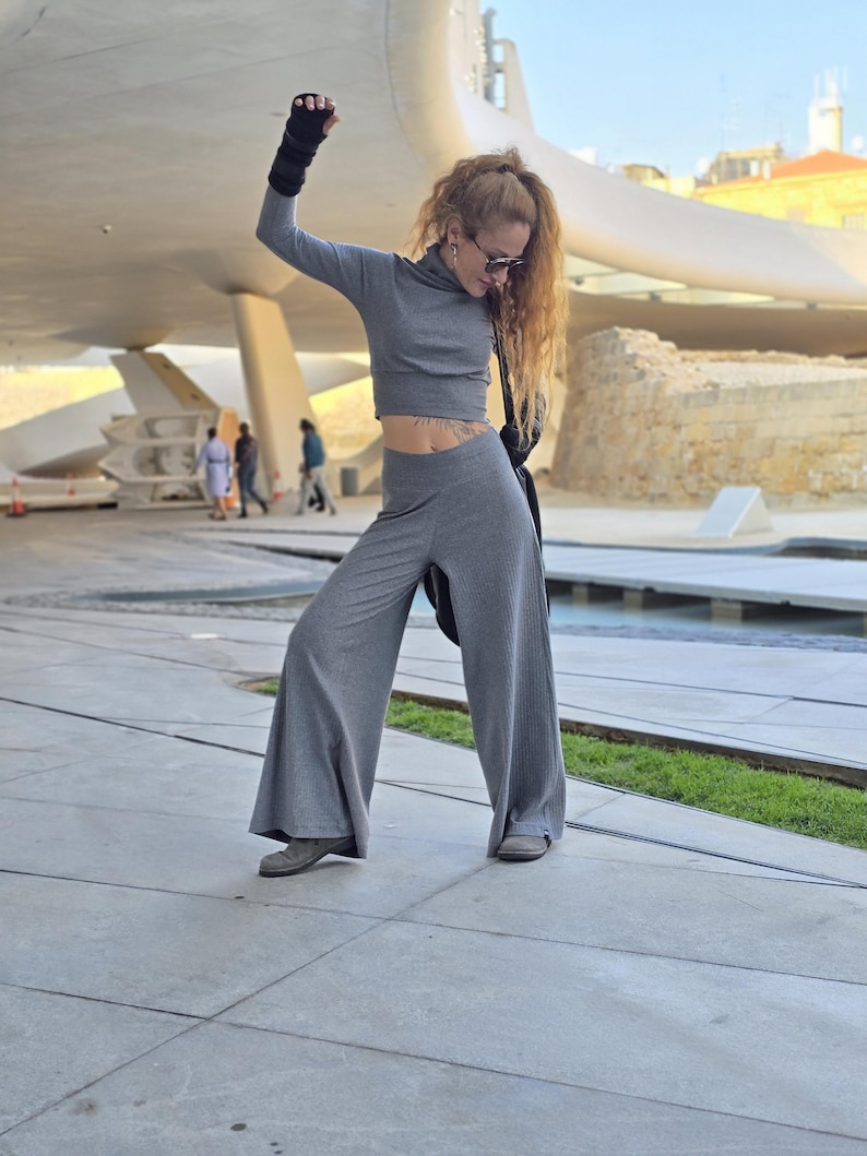 May include: A woman wearing a gray crop top and matching wide-leg pants. She is wearing black fingerless gloves and sunglasses. She is standing in front of a modern building with a curved roof.