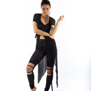 Op de afbeelding: Een vrouw draagt een zwart, doorschijnend, lang mouwig shirt met een wikkelontwerp en zwarte leggings met uitsnijdingen. De leggings hebben een hoge taille en het shirt is in de taille geknoopt.