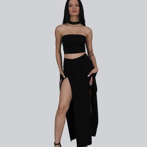 Könnte beinhalten: Ein schwarzes zweiteiliges Outfit, bestehend aus einem trägerlosen Crop-Top und einem Maxirock mit hohem Schlitz. Der Rock hat ein Wickeldesign mit Taschen. Das Model trägt schwarze Plateau-Heels und ein Halsband. Der Hintergrund ist einfarbig hellgrau.