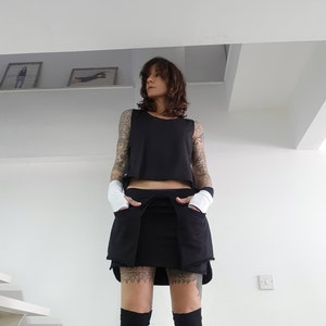May include: A woman wearing a black sleeveless top and a black skirt with pockets. She is wearing black tights and has tattoos on her legs.