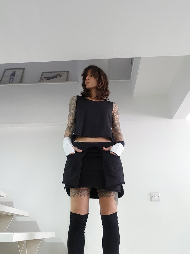 May include: A woman wearing a black sleeveless top and a black skirt with pockets. She is wearing black tights and has tattoos on her legs.