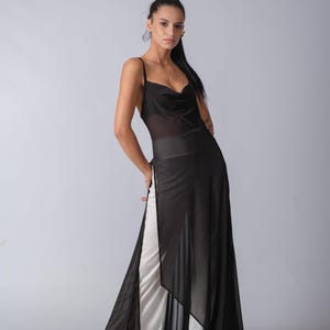 Op de afbeelding: Een vrouw draagt een zwarte, doorschijnende maxi-jurk met een cowl-hals en een witte slip eronder. De jurk heeft een zijsplit en een vloeiende silhouet.