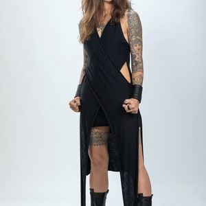 May include: A woman wearing a black, long, sleeveless dress with a thigh-high slit and a black garter belt. She has tattoos on her arms and legs and is wearing black boots.