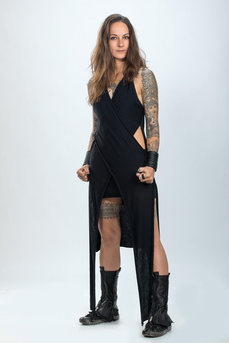 May include: A woman wearing a black, long, sleeveless dress with a thigh-high slit and a black garter belt. She has tattoos on her arms and legs and is wearing black boots.