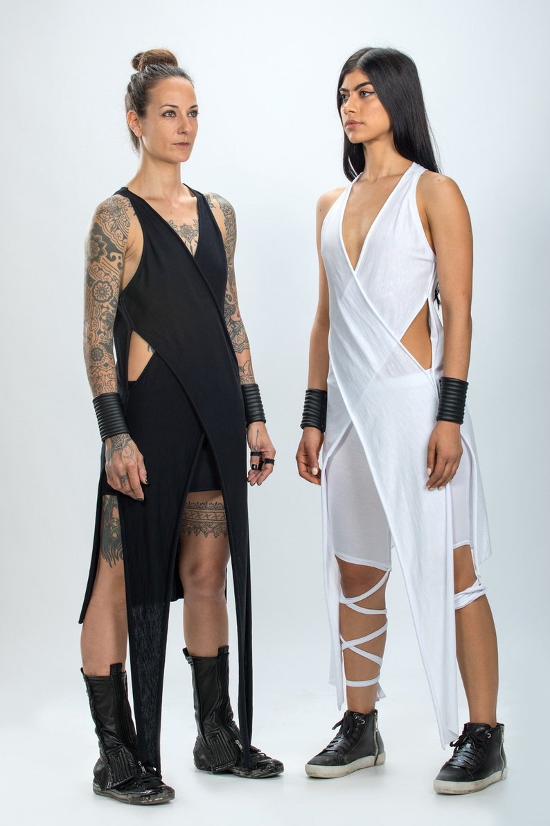 May include: Two women wearing long, asymmetrical dresses. The woman on the left is wearing a black dress with a side slit and a tattoo on her arm. The woman on the right is wearing a white dress with a side slit and a black belt.