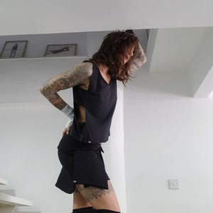 May include: A woman wearing a black crop top with a side cut-out and a black skirt with a layered design. She has tattoos on her arms and legs.