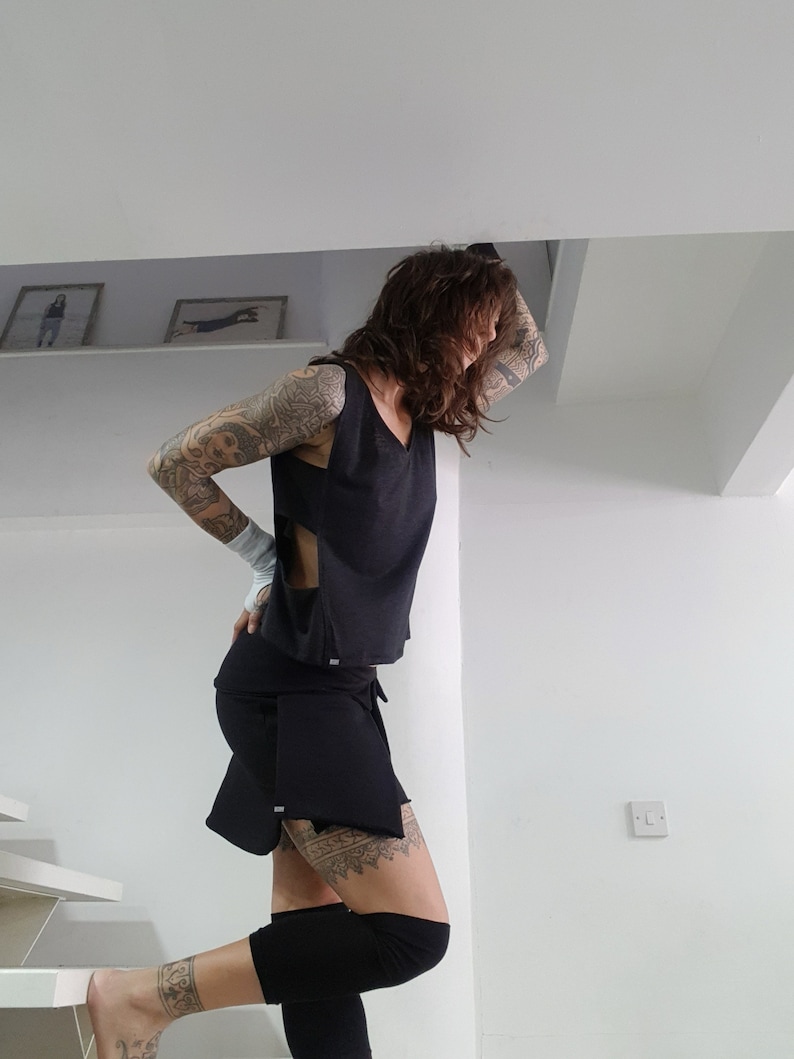 May include: A woman wearing a black crop top with a side cut-out and a black skirt with a layered design. She has tattoos on her arms and legs.
