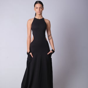 May include: A woman wearing a black maxi dress with a cut-out design on the sides. The dress has a halter neckline and a flowing skirt. She is wearing black sandals.