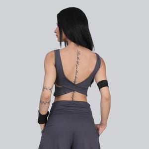 May include: A dark gray crop top and matching pants. The top has a crisscross design in the back. The model is wearing a black wristband on the left arm. The back of the model has a tattoo.