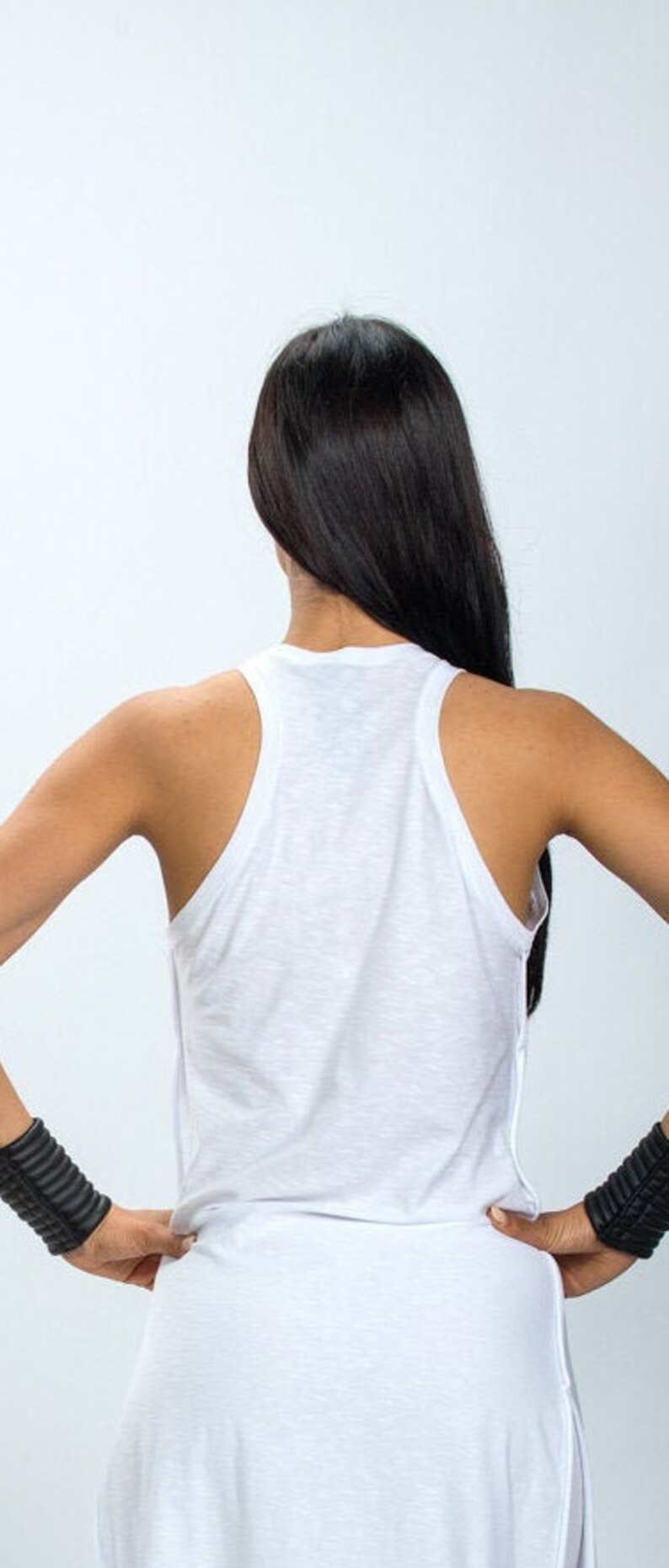 May include: A white sleeveless tank top with a long, flowing design. The tank top has a relaxed fit and a slightly loose neckline. The model is wearing the tank top with black wristbands.
