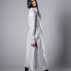 May include: A white long-sleeved tunic with a high collar and side slits. The tunic is made of a soft, lightweight fabric and has a relaxed fit. The model is wearing black pants and black boots.