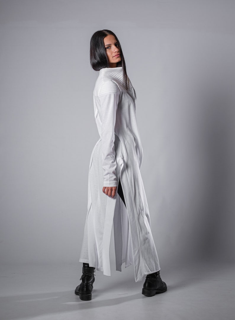 May include: A white long-sleeved tunic with a high collar and side slits. The tunic is made of a soft, lightweight fabric and has a relaxed fit. The model is wearing black pants and black boots.