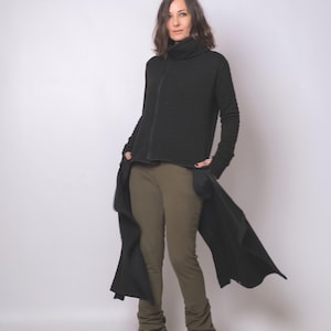 Peut inclure: Longue veste noire à manches longues avec un col montant et une fermeture éclair sur le devant. La veste a de longs panneaux fluides de chaque côté. Le modèle porte un pantalon vert olive et des baskets grises avec des touches de rouge.