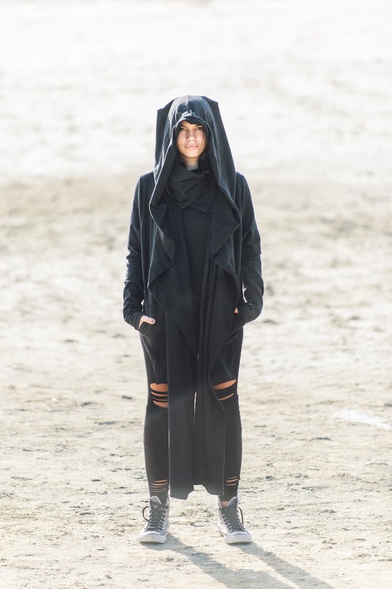 May include: A person wearing a black hooded cloak with a long, flowing design. The cloak has a high collar and a wide hood that covers the person's head. The person is standing on a sandy surface.