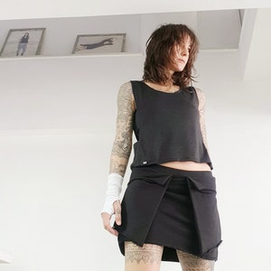 May include: A woman wearing a black sleeveless top, a black skirt, and black knee-high socks. She has a tattoo on her arm and leg.