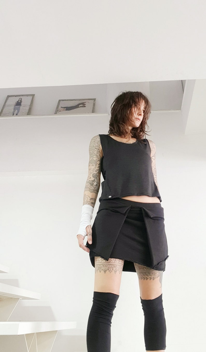 May include: A woman wearing a black sleeveless top, a black skirt, and black knee-high socks. She has a tattoo on her arm and leg.