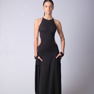 May include: A woman wearing a black maxi dress with a high neckline and cutouts on the sides. She is standing in front of a white background.