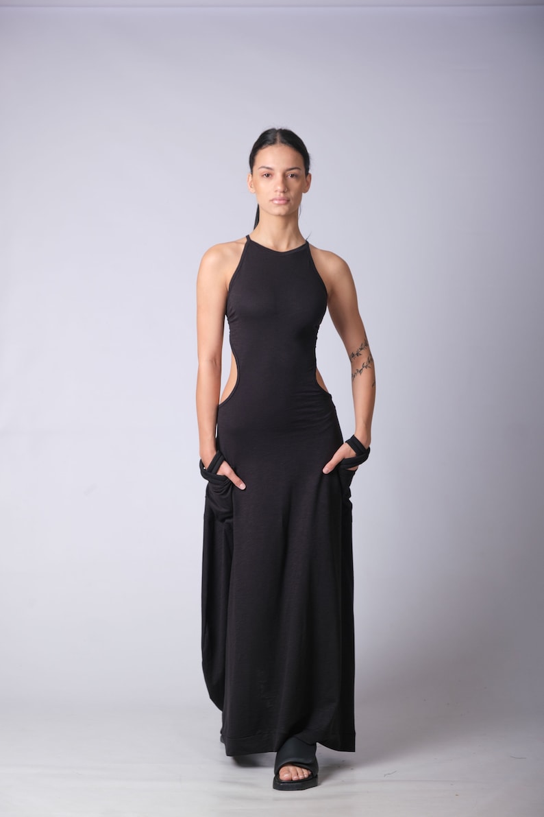May include: A woman wearing a black maxi dress with a high neckline and cutouts on the sides. She is standing in front of a white background.