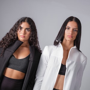 May include: Two women wearing black and white jackets with a similar style. The woman on the left has curly hair and is wearing a black jacket with a high collar. The woman on the right has straight hair and is wearing a white jacket with a high collar.
