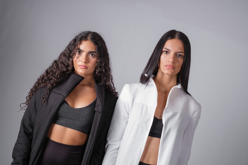 May include: Two women wearing black and white jackets with a similar style. The woman on the left has curly hair and is wearing a black jacket with a high collar. The woman on the right has straight hair and is wearing a white jacket with a high collar.