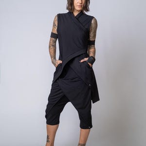 Könnte beinhalten: Eine schwarze ärmellose Tunika mit Wickeloptik, Stehkragen und Seitentaschen, kombiniert mit einer schwarzen, wadenlangen Hose mit tiefem Schritt. Das Outfit wird durch schwarze Pantoletten mit breiten, gepolsterten Riemen ergänzt.