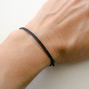 Puede incluir: Un simple brazalete de cuerda negra usado en una muñeca.