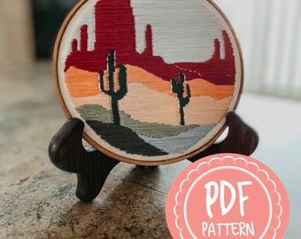 Può includere: Arte di ricamo a cerchio che rappresenta un paesaggio desertico con formazioni rocciose rosse, cactus e un cielo blu. Il cerchio è su un supporto in legno e un cerchio rosa con il testo "PDF PATTERN" è in primo piano.
