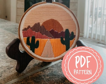 Può includere: Un modello PDF per un progetto di ricamo su un telaio che rappresenta un paesaggio desertico con montagne, una strada, un tramonto e due cactus. Il telaio è su un supporto in legno.