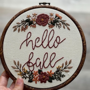 May include: Embroidered hoop art with the text "hello fall" in cursive script. The hoop is decorated with a floral wreath of burgundy, orange, and green flowers.