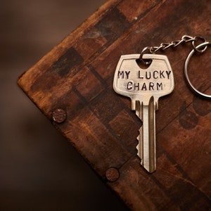 Puede incluir: Un llavero plateado con forma de llave con las palabras "MY LUCKY CHARM" grabadas. La llave está unida a una cadena y un anillo. El llavero descansa sobre una superficie de madera marrón oscura y envejecida.