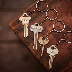 May include: Four keychains with different words on them: "LOVE", "NASHVILLE", "BRAVE", and "STRONG". The keychains are on a wooden surface.