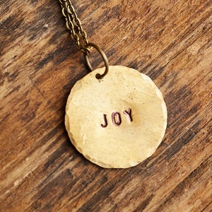 Puede incluir: Un collar de oro con un colgante redondo que dice "JOY" en letras rojas.